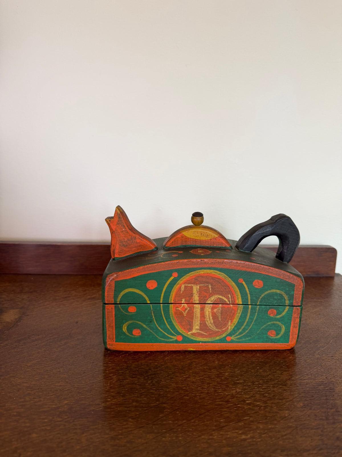 A small hand-painted wooden keepsake box with a lid, featuring a green and orange design with a central monogram 'TC' and decorative scrollwork, resting on a dark wooden table against a plain off-white wall.