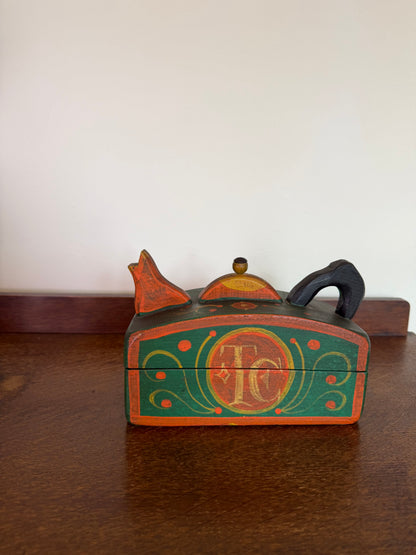 A small hand-painted wooden keepsake box with a lid, featuring a green and orange design with a central monogram 'TC' and decorative scrollwork, resting on a dark wooden table against a plain off-white wall.