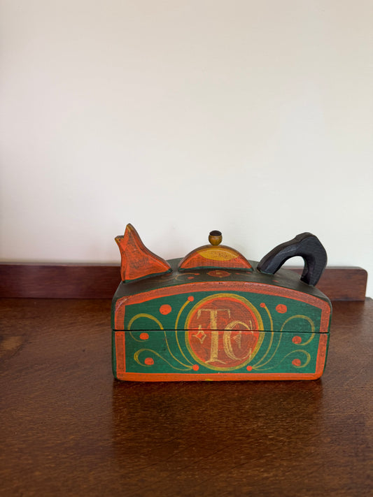A small hand-painted wooden keepsake box with a lid, featuring a green and orange design with a central monogram 'TC' and decorative scrollwork, resting on a dark wooden table against a plain off-white wall.