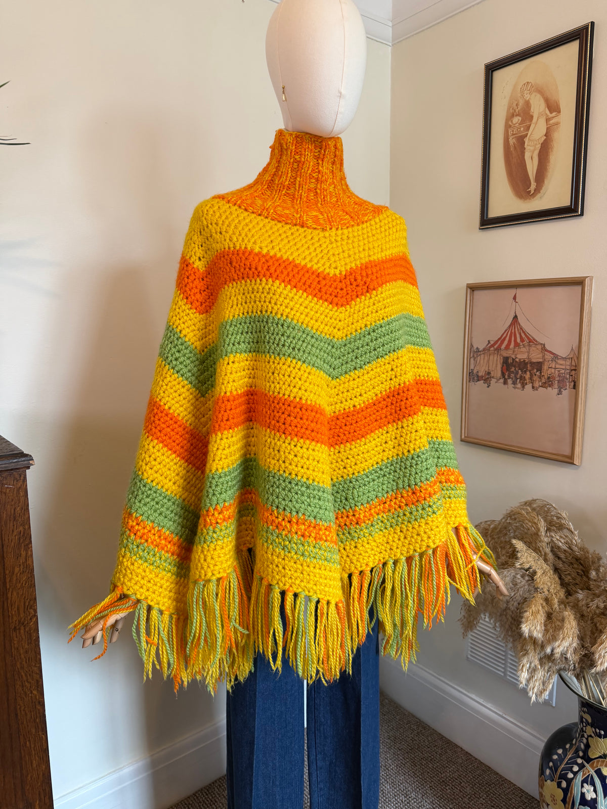 A crocheted poncho sweater with horizontal stripes of yellow, orange, and green, featuring a mock neck and fringe trim, displayed on a mannequin in an indoor room setting.
