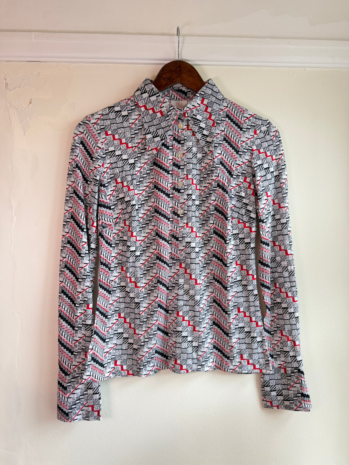 A long-sleeve vintage collared button-up shirt with a geometric zigzag pattern in black, white, and red, hanging on a wooden hanger against a plain off-white wall.