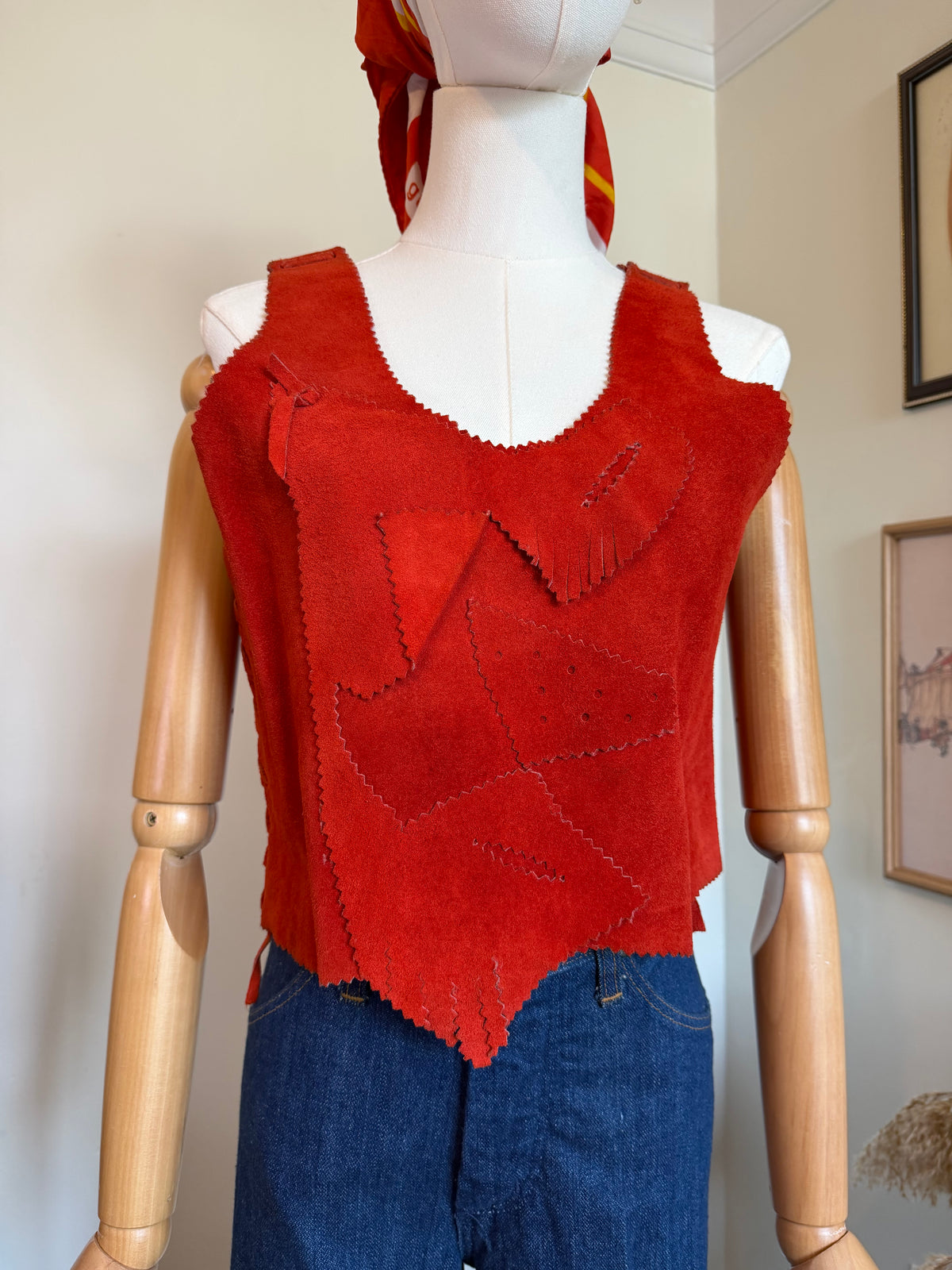 A red suede vest with geometric patchwork panels and fringe details, displayed on a wooden mannequin torso wearing blue jeans. The mannequin is positioned indoors against a light beige wall.