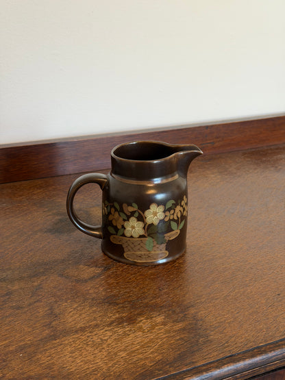 Vintage Royal Doulton Lambeth Stoneware milk jug in the Basque pattern, featuring a dark brown glaze with cream and green floral basket motif and curved handle, photographed on a wooden surface.