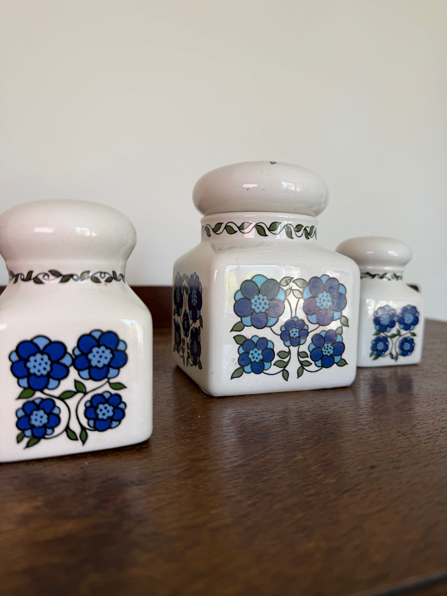 Set of three vintage Taunton Vale Pottery ceramic canisters in cream glaze with bold blue floral designs and rounded lids, photographed on a wooden surface. One large square storage jar is accompanied by two smaller matching shakers, with maker’s mark visible on the underside.