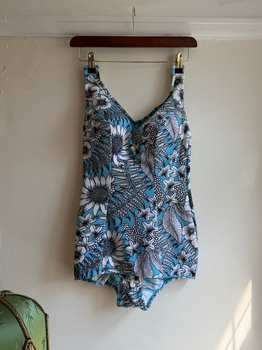 A sleeveless V-neck top with a blue, gray, and white floral and leaf pattern, hanging on a clear hanger with a brown wooden clip against an off-white wall. Vintage 1970s blue, white and black tropical floral one-piece swimsuit with oversized botanical print and V-neckline.