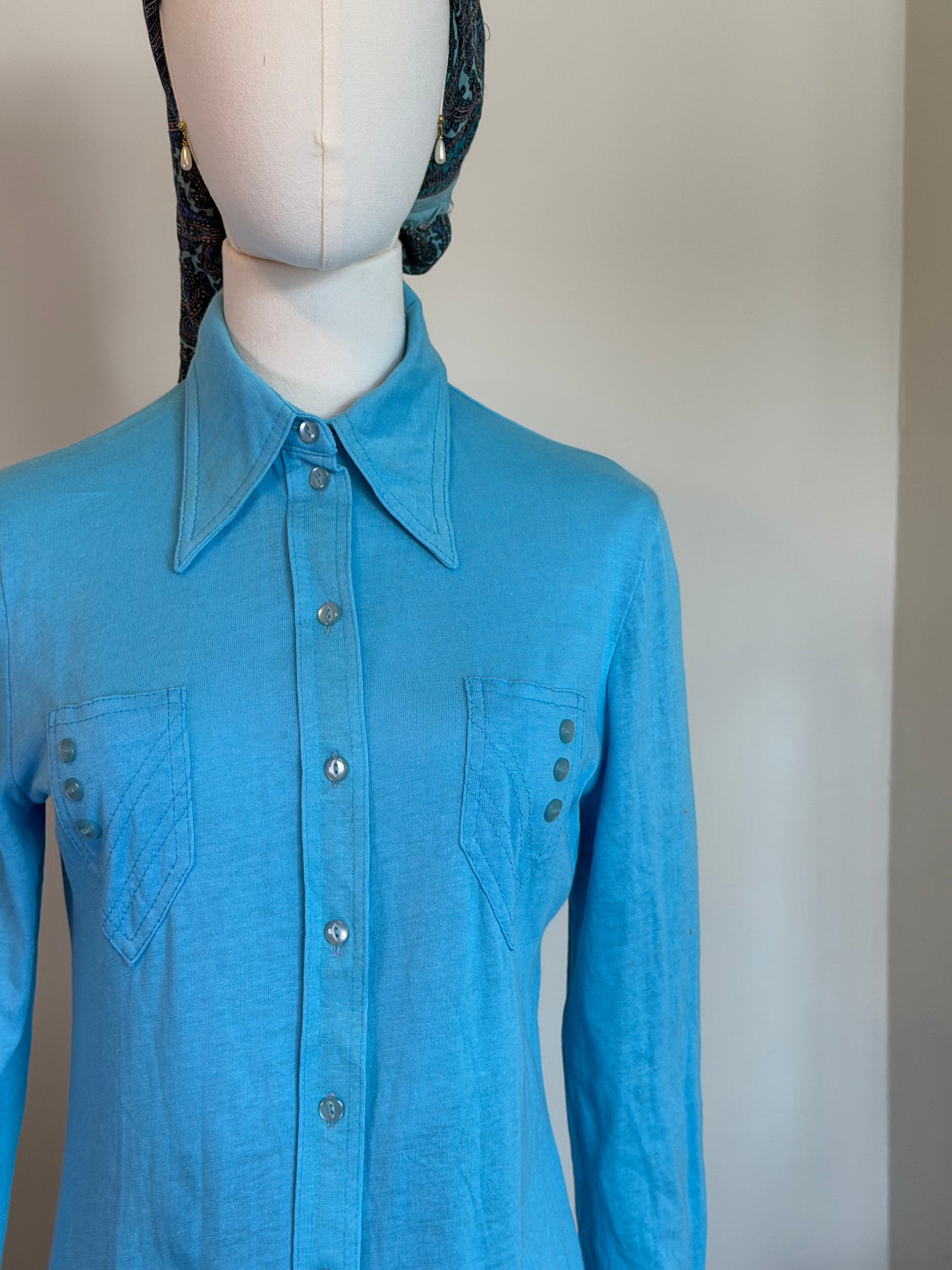 A bright blue long-sleeve button-up shirt with a pointed collar and two chest pockets, displayed on a white mannequin torso against a plain light wall.