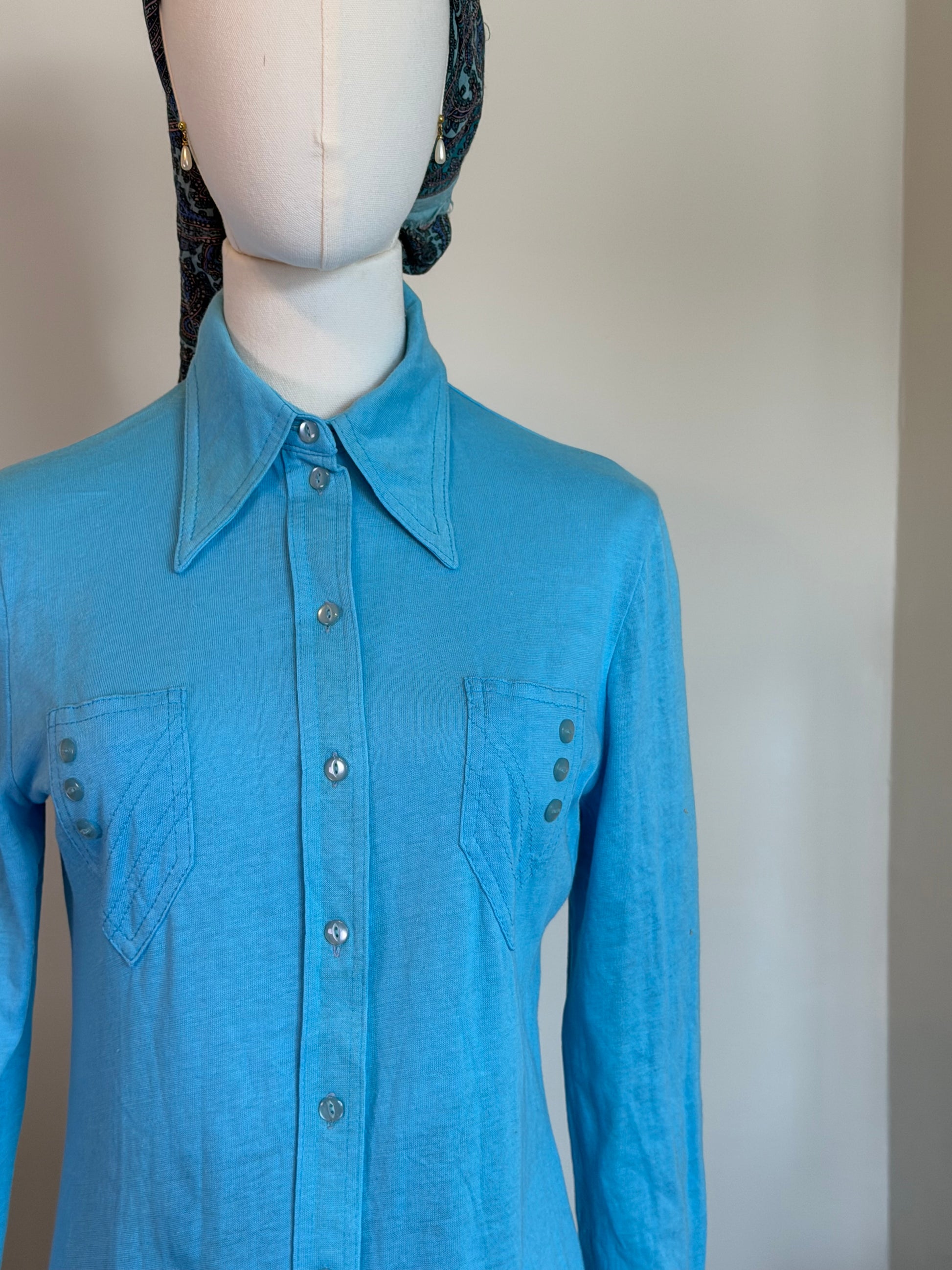 A bright blue long-sleeve button-up shirt with a pointed collar and two chest pockets, displayed on a white mannequin torso against a plain light wall.
