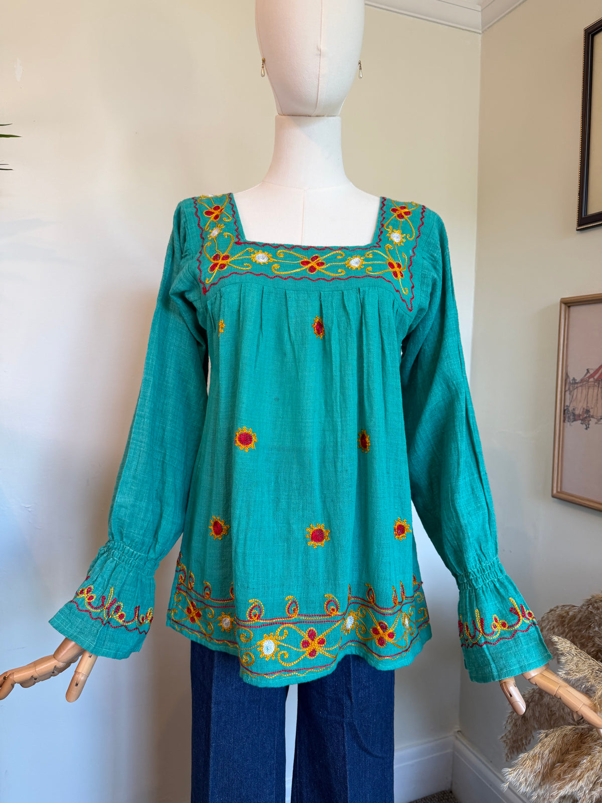 Vintage 1970s turquoise cotton smock top with colourful floral embroidery around the square neckline, hem and flared cuffs, styled on a mannequin with blue jeans.