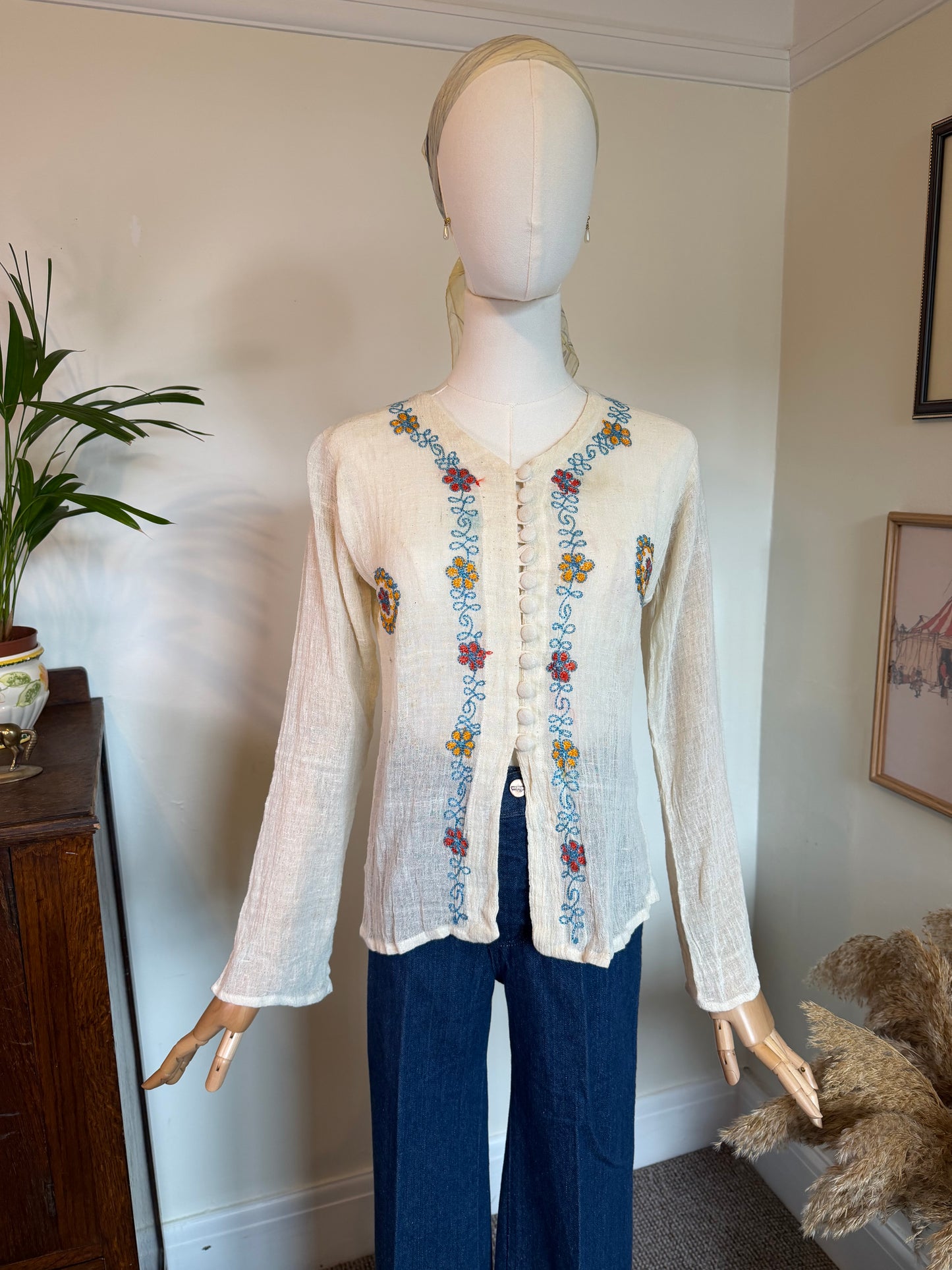 A cream-colored, long-sleeve linen cardigan with colorful floral embroidery down the front and on the pockets, displayed on a mannequin with dark blue trousers in an indoor room setting.