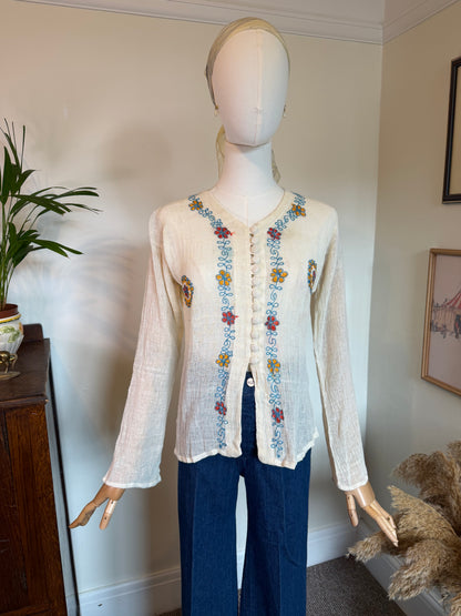 A cream-colored, long-sleeve linen cardigan with colorful floral embroidery down the front and on the pockets, displayed on a mannequin with dark blue trousers in an indoor room setting.