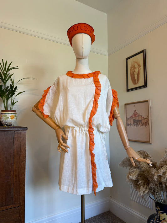 A white linen tunic dress with bright orange ruffle trim and a matching orange headband displayed on a wooden mannequin in a home interior setting.