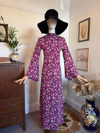 Vintage 1970s maxi dress in a rich purple and magenta floral print with a high neckline, dramatic bell sleeves and a slim column silhouette, displayed on a mannequin wearing a black floppy hat. The dress features an all-over white daisy-style flower pattern and falls to a full floor-length hem.
