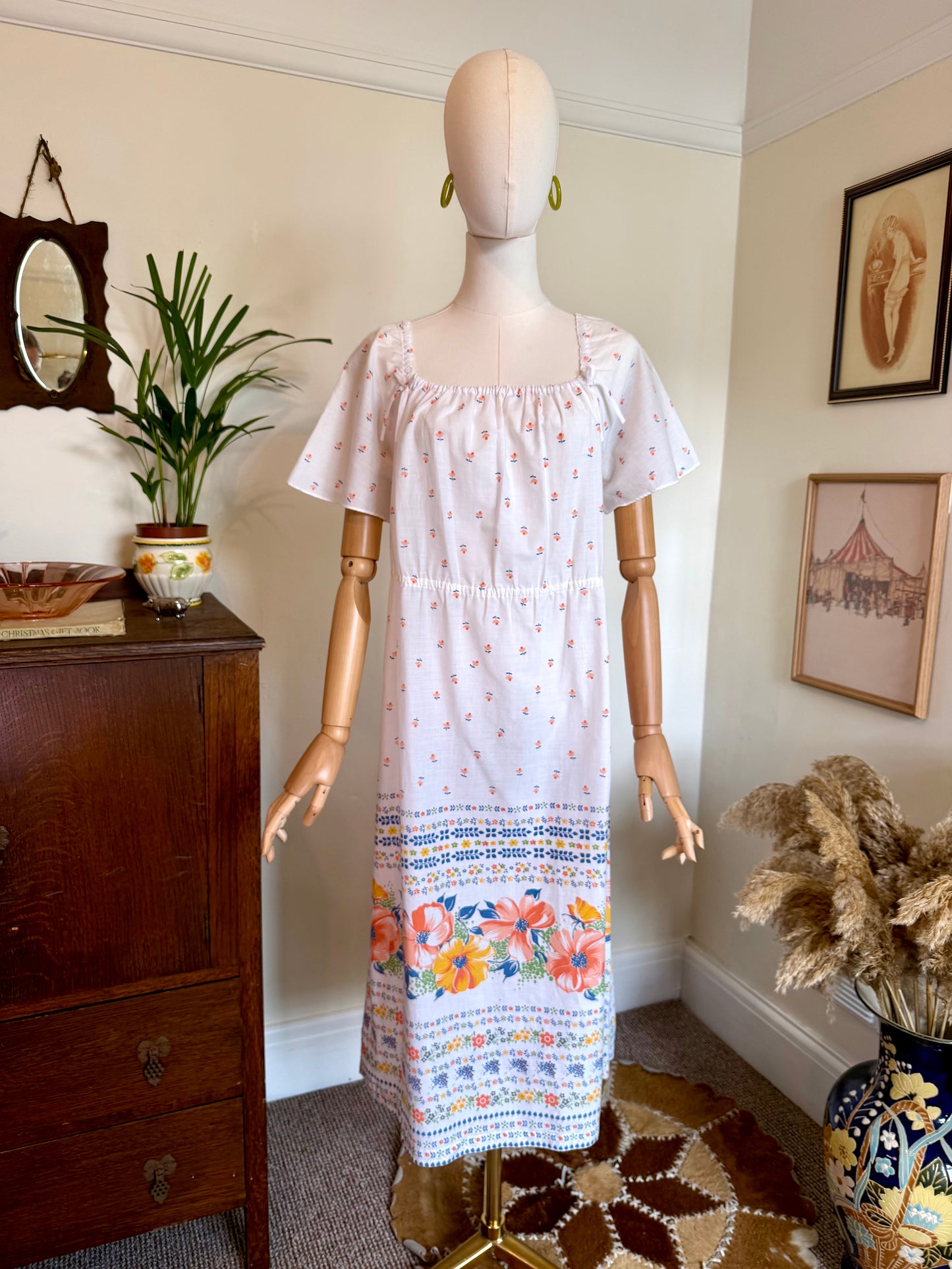 Vintage 1970s white floral maxi dress with flutter sleeves, a square gathered neckline, elasticated empire waist and colourful border print featuring coral, peach and yellow flowers around the hem, displayed on a mannequin with green hoop earrings.