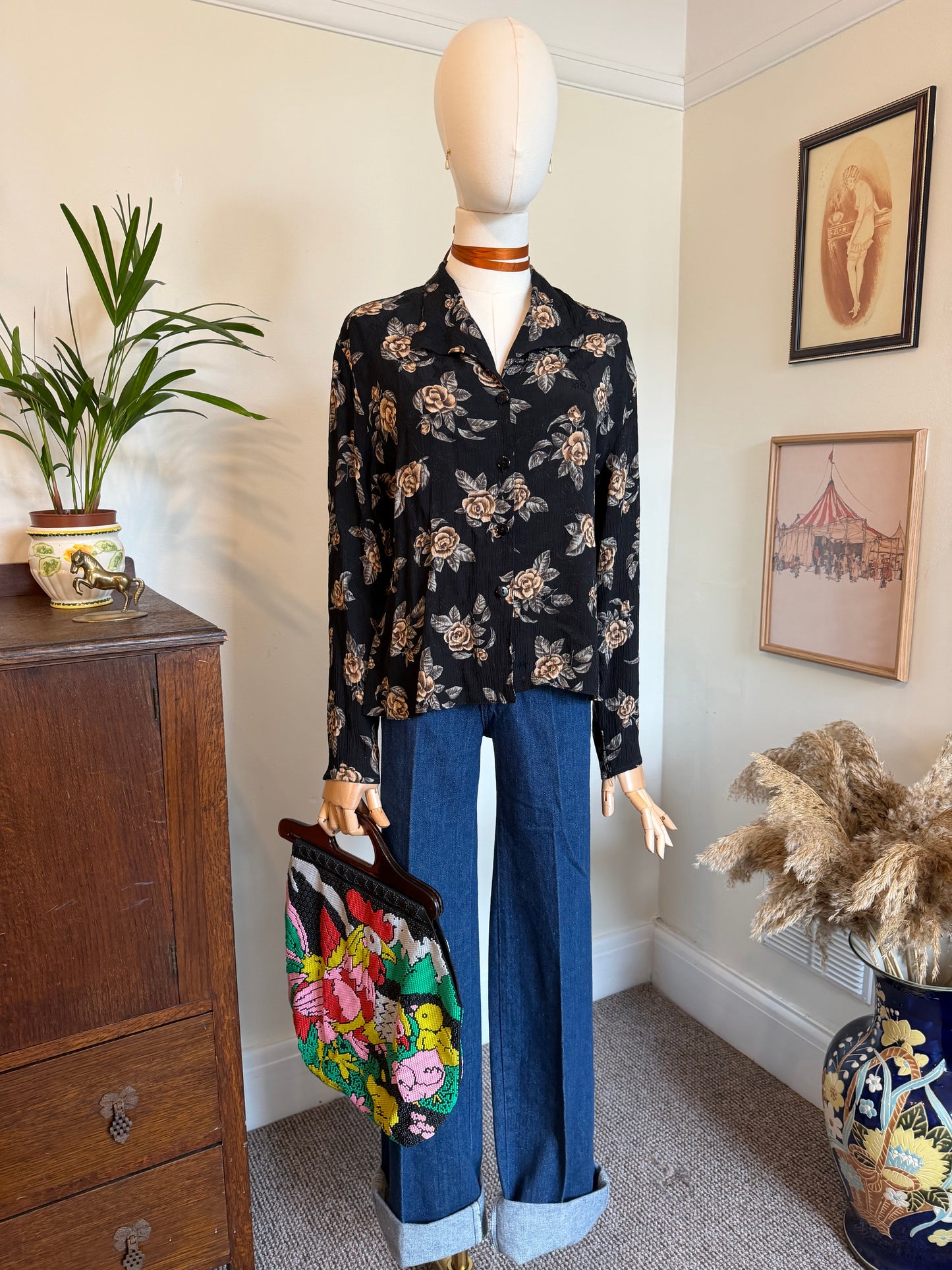 A vintage-style outfit displayed on a mannequin in a home interior, featuring a black long-sleeve button-up blouse with a beige and brown floral print, paired with wide-leg blue jeans with rolled cuffs, and holding a colorful floral handbag.