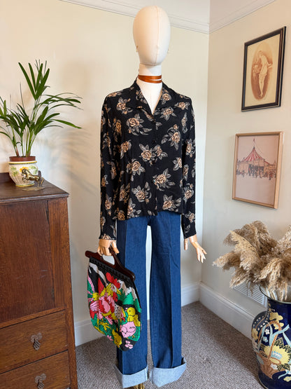 A vintage-style outfit displayed on a mannequin in a home interior, featuring a black long-sleeve button-up blouse with a beige and brown floral print, paired with wide-leg blue jeans with rolled cuffs, and holding a colorful floral handbag.
