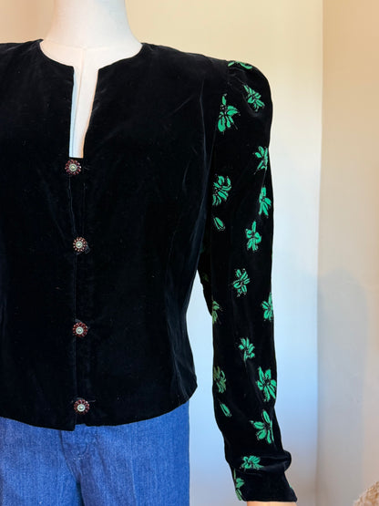 A black velvet long-sleeve blouse with green floral embroidery on the sleeves, displayed on a mannequin with blue jeans against a plain beige wall.
