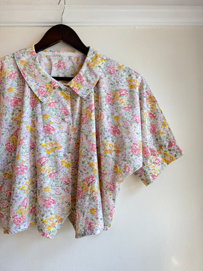 Vintage floral short-sleeve blouse with an oversized pointed collar, featuring pink roses, yellow and lilac flowers and green foliage on a pale background.