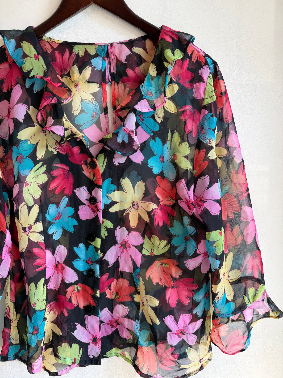 Vintage colourful floral sheer blouse with a black base, oversized pink, turquoise, red, green and yellow flowers, ruffled neckline and long flared sleeves. A sheer black blouse with a vibrant, multicolored floral print, hanging on a dark wooden hanger against a plain off-white wall.