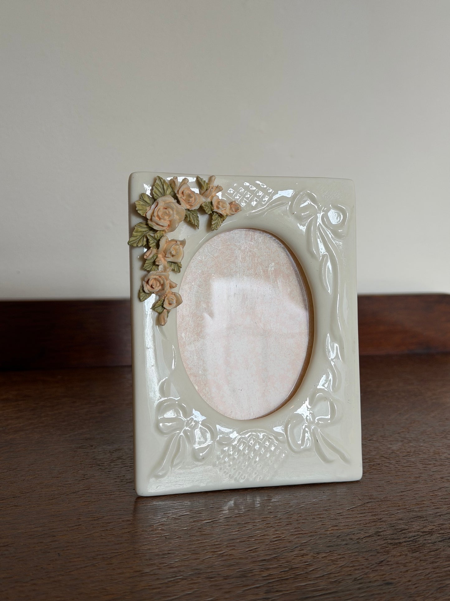 A square cream-colored ceramic picture frame with embossed floral and scroll details, featuring a small cluster of 3D roses in beige and green on the bottom-left corner, set against a split background of a dark wood surface and a plain light wall.