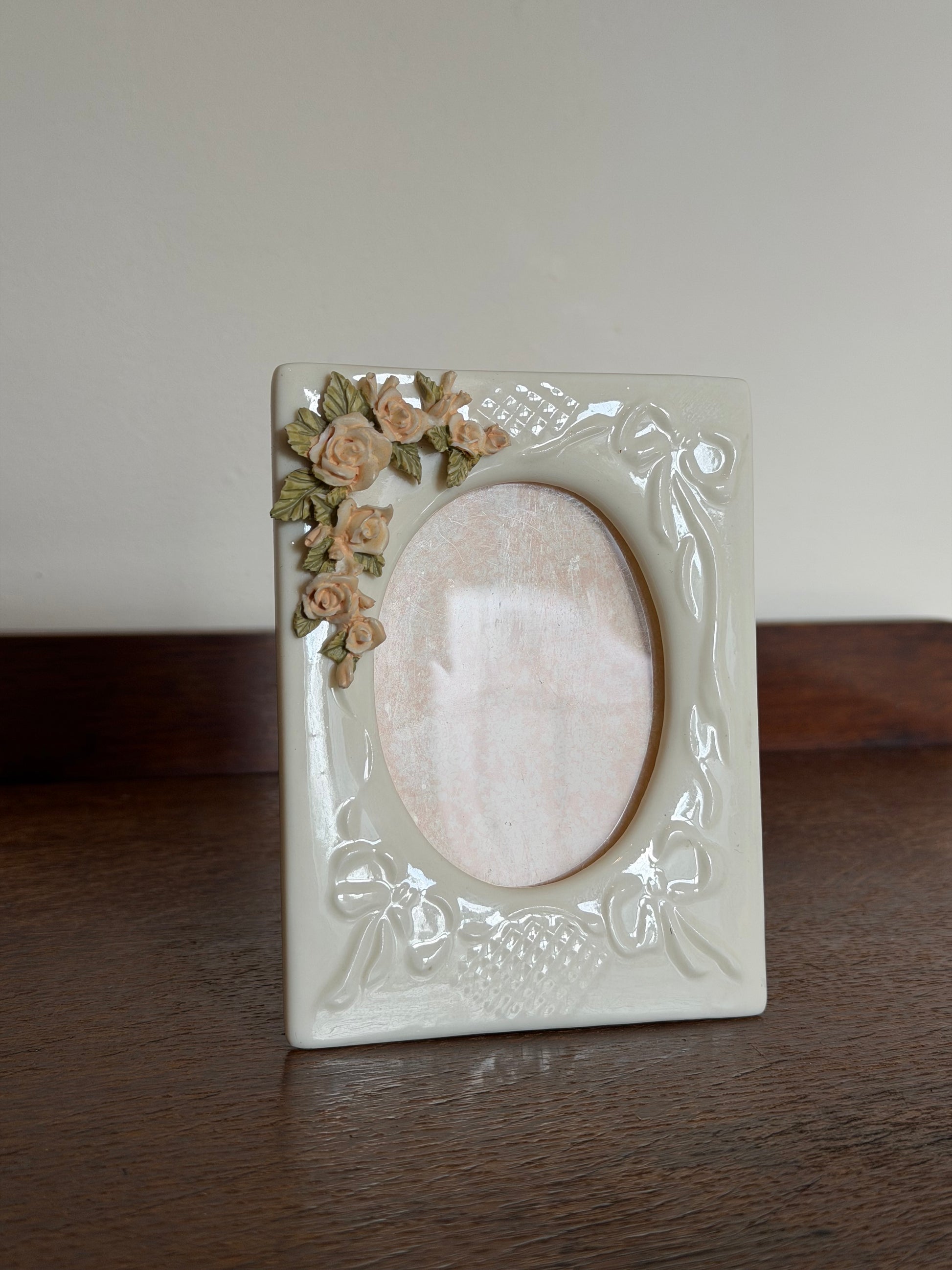 A square cream-colored ceramic picture frame with embossed floral and scroll details, featuring a small cluster of 3D roses in beige and green on the bottom-left corner, set against a split background of a dark wood surface and a plain light wall.