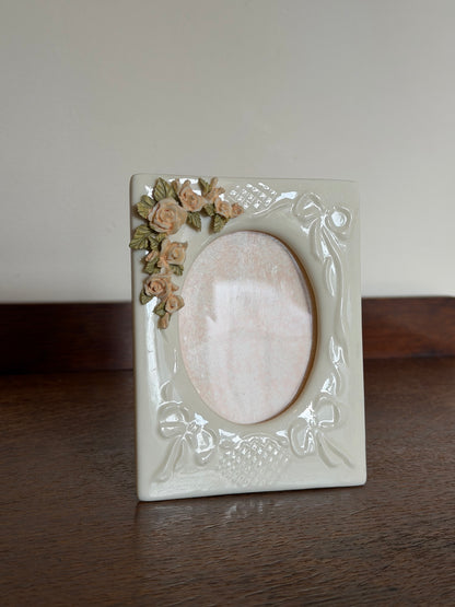 A square cream-colored ceramic picture frame with embossed floral and scroll details, featuring a small cluster of 3D roses in beige and green on the bottom-left corner, set against a split background of a dark wood surface and a plain light wall.