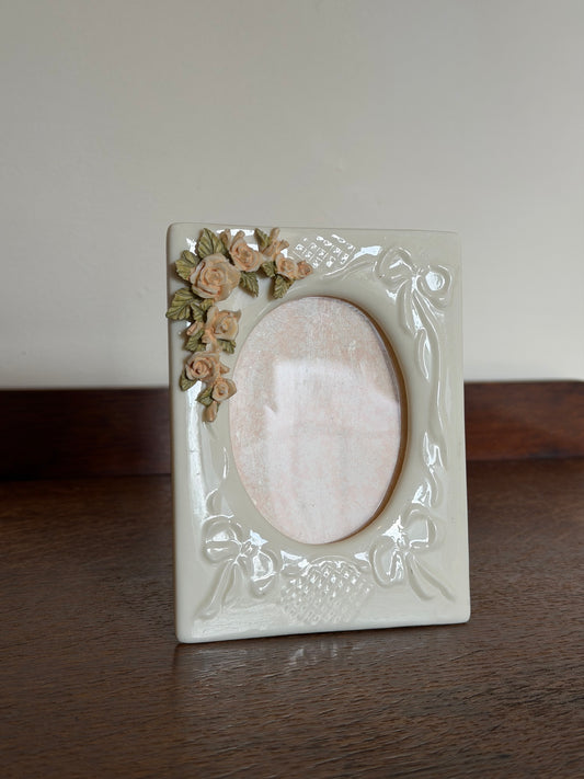 A square cream-colored ceramic picture frame with embossed floral and scroll details, featuring a small cluster of 3D roses in beige and green on the bottom-left corner, set against a split background of a dark wood surface and a plain light wall.