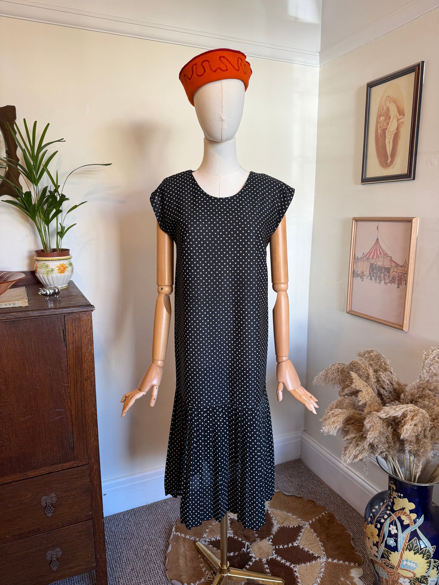 Vintage 1980s black midi dress with a small white polka dot print, cap sleeves, scoop neckline and gathered drop-waist flounce hem, displayed on a mannequin wearing a bright orange hat.
