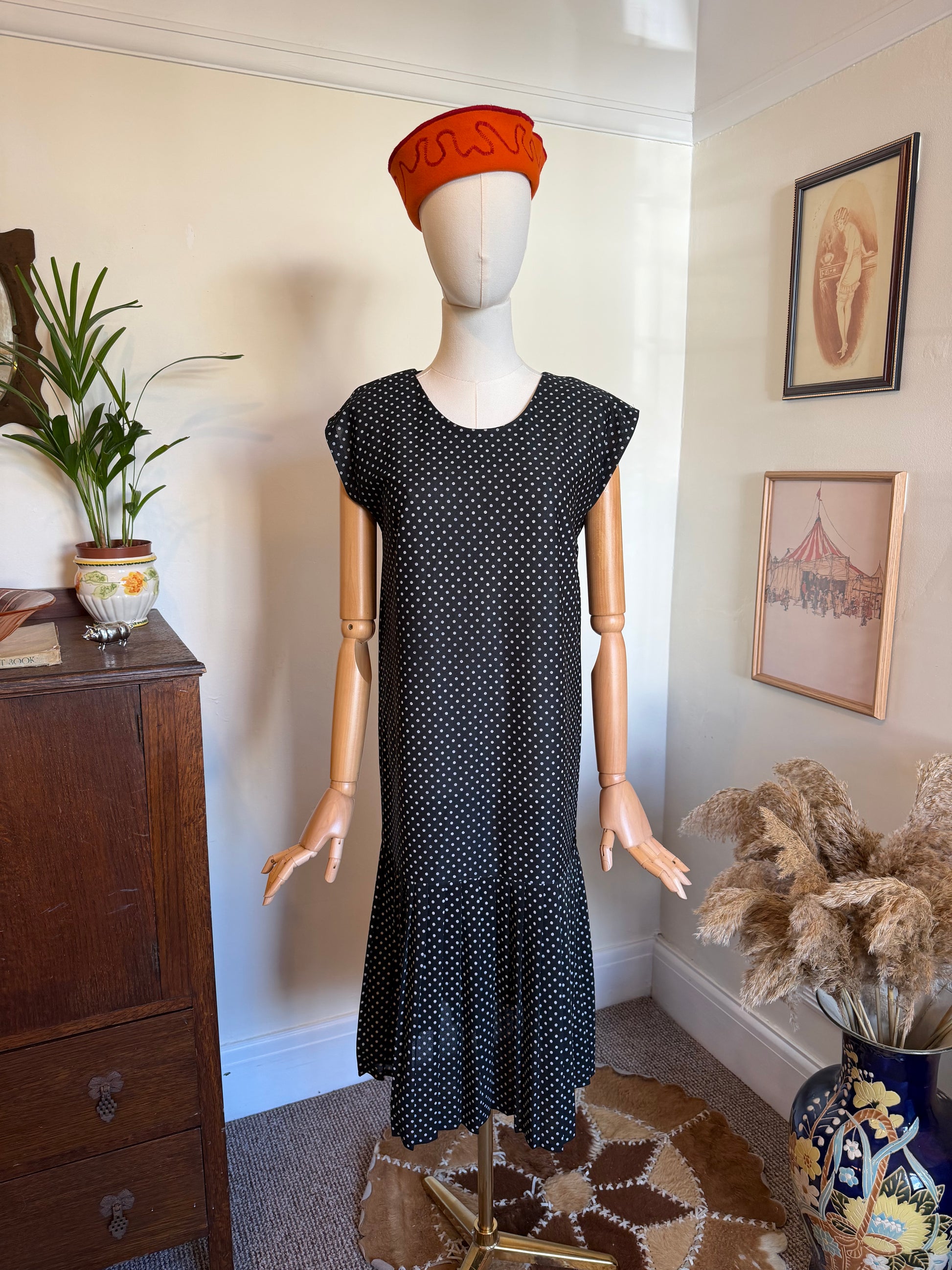 Vintage 1980s black midi dress with a small white polka dot print, cap sleeves, scoop neckline and gathered drop-waist flounce hem, displayed on a mannequin wearing a bright orange hat.
