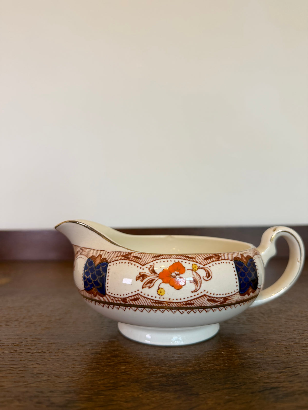 A vintage ceramic gravy boat with a cream-colored body, decorated with a brown floral border and blue and orange floral motifs, resting on a dark wooden table against a plain beige wall.