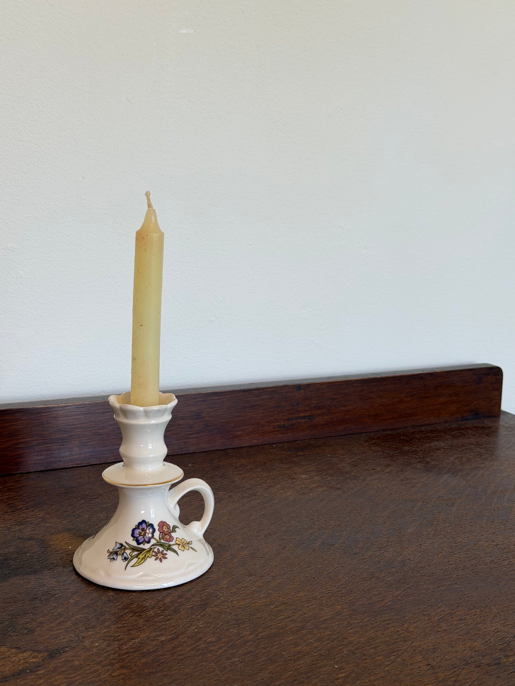 Vintage Belleek Galway Irish porcelain candle holder with a loop handle and scalloped top, decorated with delicate blue, pink and yellow wildflowers and subtle gold trim. Photographed with a beeswax taper candle against an off-white wall on a dark wooden surface.