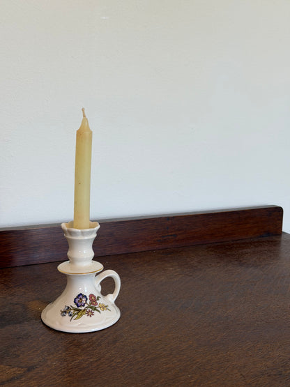 Vintage Belleek Galway Irish porcelain candle holder with a loop handle and scalloped top, decorated with delicate blue, pink and yellow wildflowers and subtle gold trim. Photographed with a beeswax taper candle against an off-white wall on a dark wooden surface.