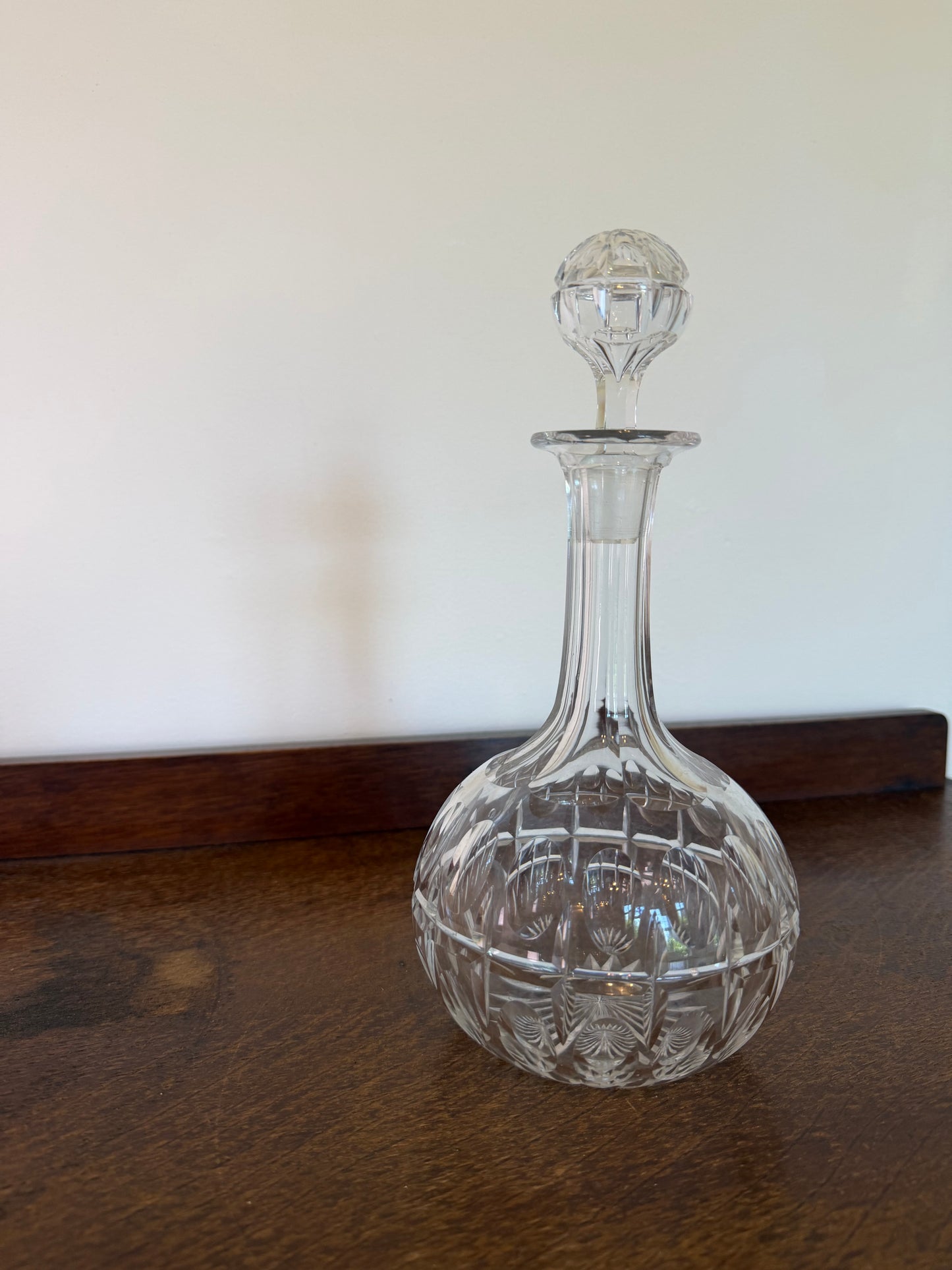 A clear cut-crystal decanter with a matching stopper, displayed on a dark wooden surface against a plain off-white background.