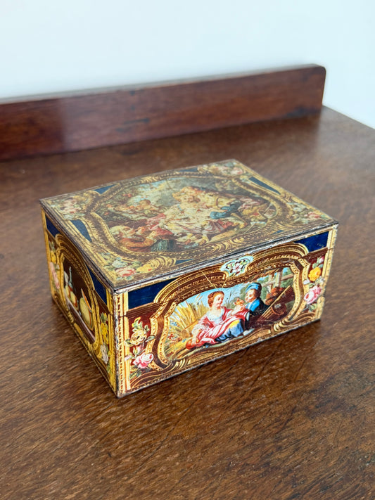 A small rectangular decorative box with ornate classical-style painted illustrations and gold detailing, placed on a dark wooden table. Vintage decorative tin keepsake box with Rococo-inspired artwork, hinged lid and gold interior displayed on a wooden sideboard.