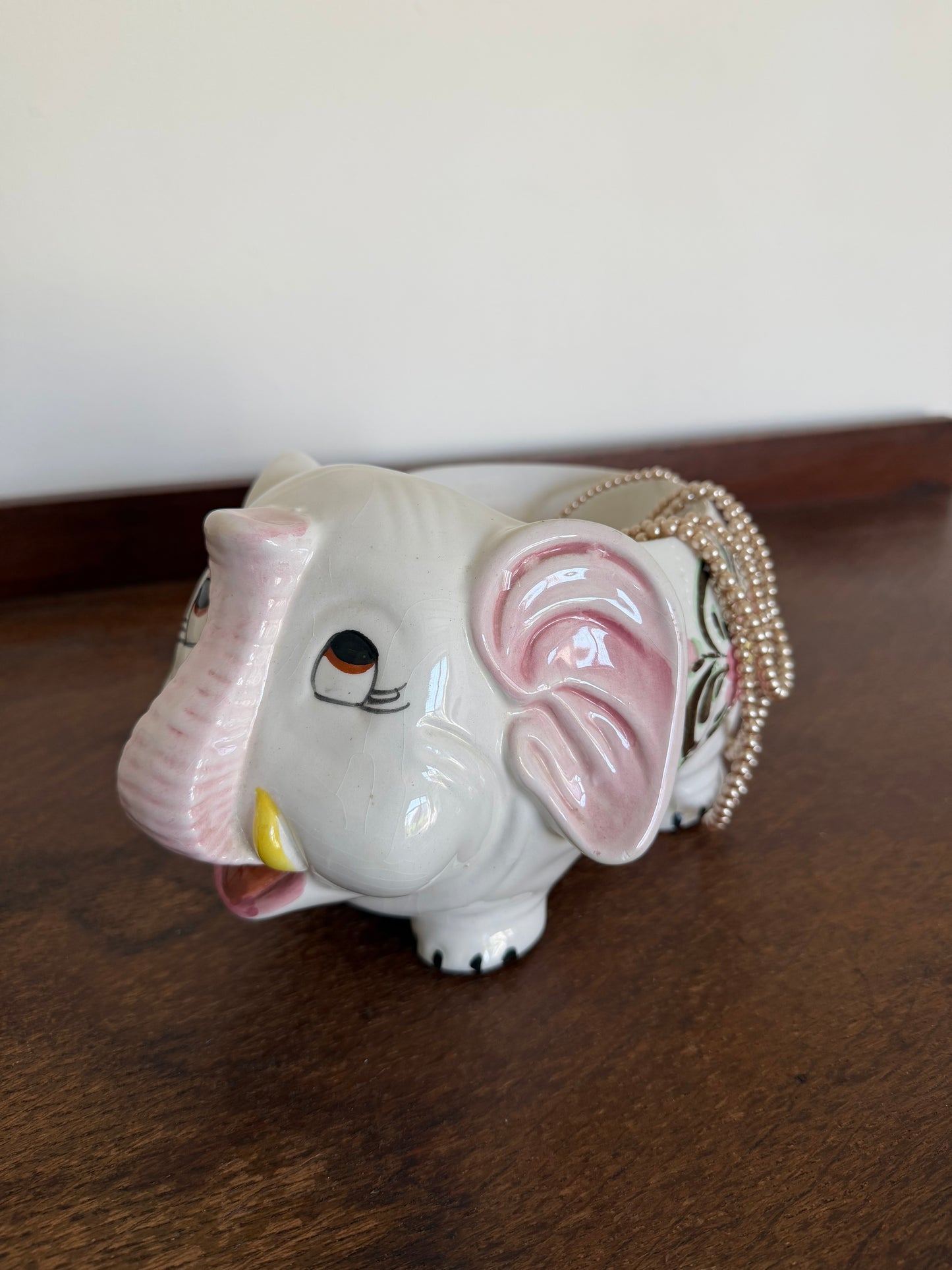 Vintage ceramic elephant planter with a glossy white finish, pink ears and trunk details, and hand-painted pink flowers with brown foliage. The elephant-shaped planter stands on four short legs and is photographed against a warm wooden background. Styled with a string of draped pearls 