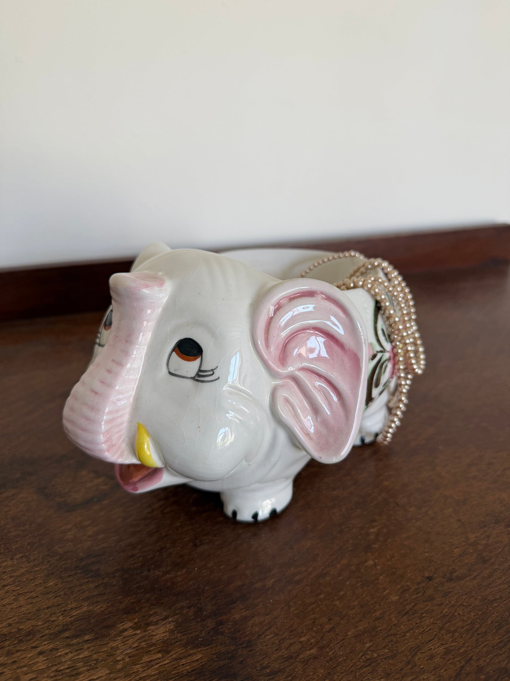 Vintage ceramic elephant planter with a glossy white finish, pink ears and trunk details, and hand-painted pink flowers with brown foliage. The elephant-shaped planter stands on four short legs and is photographed against a warm wooden background. Styled with a string of draped pearls 