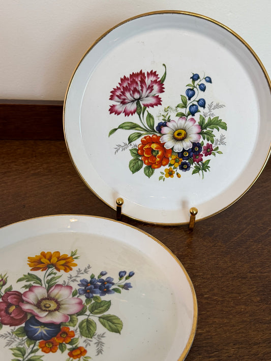 Two vintage floral ceramic plates with gold trim, displayed on a wooden surface with a wall in the background.