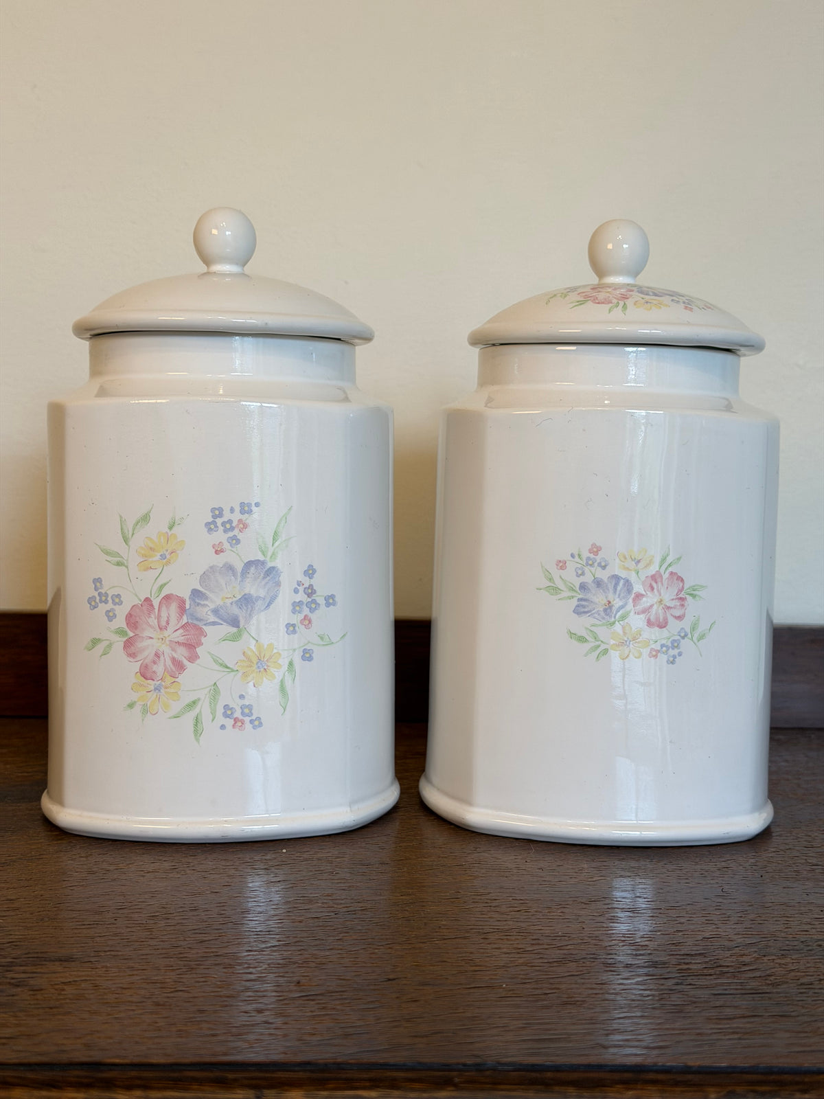 Pair of vintage ivory ceramic storage jars with matching lids and pastel pink, blue and yellow floral bouquets, displayed together on a wooden surface.