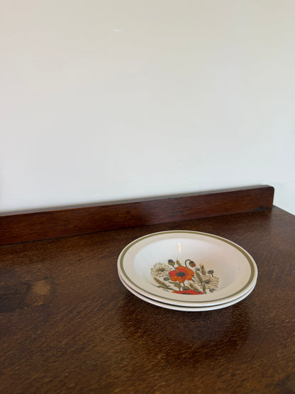 A small round ceramic plate with a floral poppy design in red, blue, and green, resting on a dark wooden table against an off-white wall. 
Pair of vintage J&G Meakin Ironstone bowls with orange poppy floral pattern on cream china.

