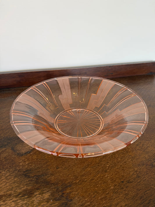 A clear glass bowl with a warm pinkish tint and radial ribbed detailing, shown resting on a dark brown wooden surface next to a white wall. 1930s pink pressed glass Art Deco fruit bowl with geometric sunburst pattern.