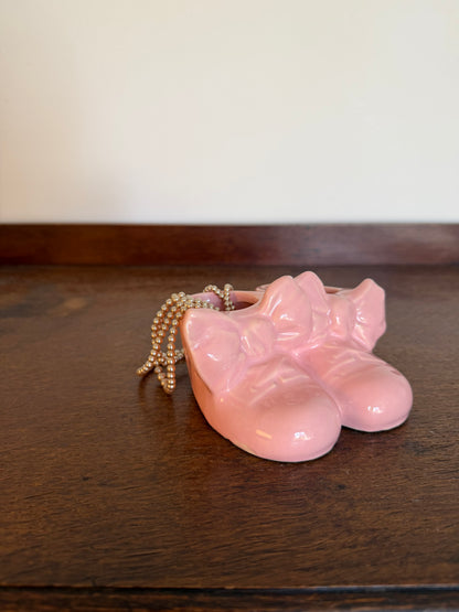 Vintage ceramic planter shaped like a pair of baby booties, finished in glossy pale pink with large bows across the front. The joined shoes have open tops for planting and are photographed from multiple angles showing the sides, top and base. Styled with a draped string of pearls 