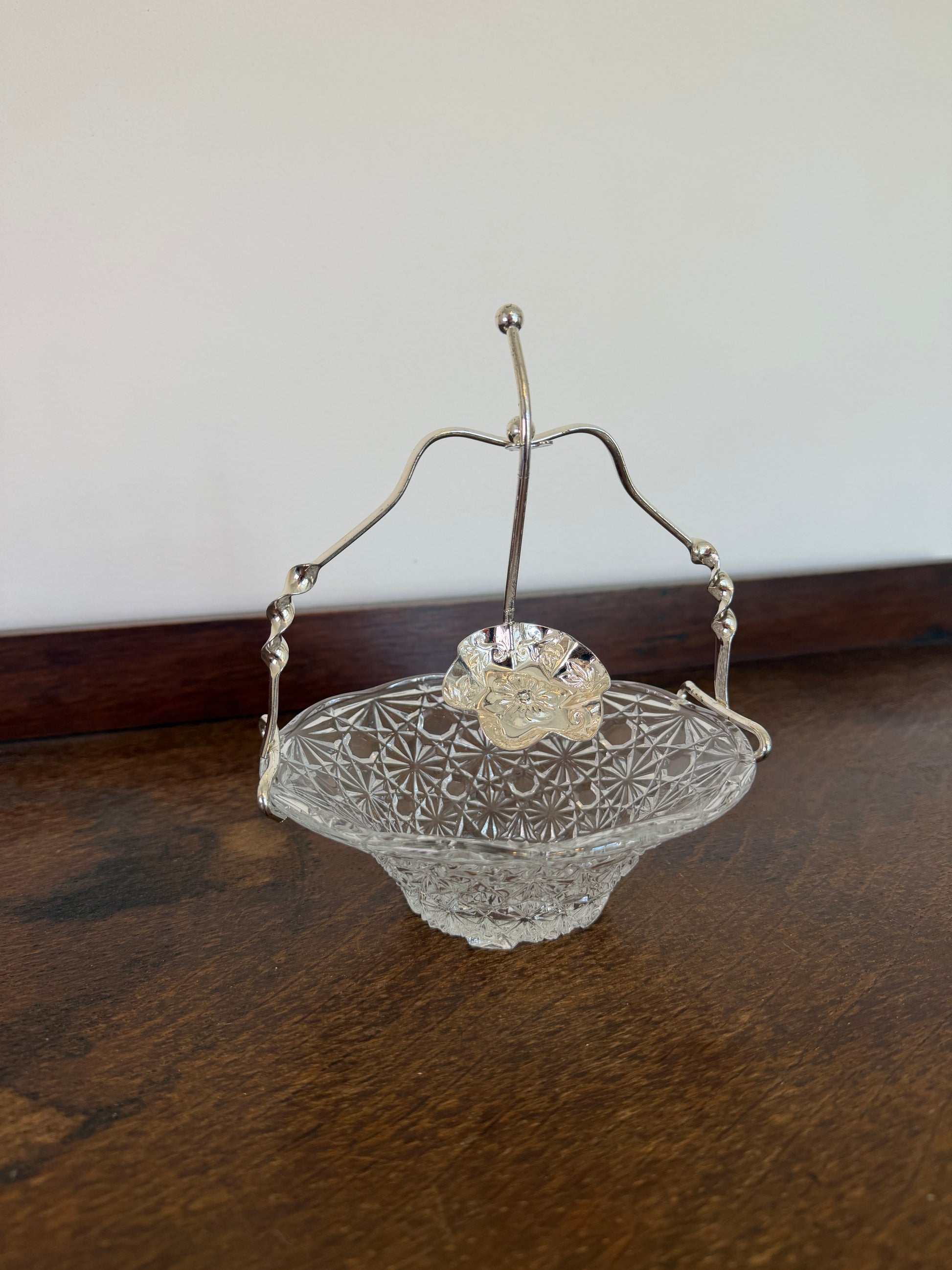 A vintage clear glass candy bowl with a cut star pattern, featuring a silver-tone metal handle and a small clear glass flower accent on top, resting on a dark wooden table.