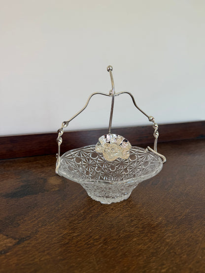 A vintage clear glass candy bowl with a cut star pattern, featuring a silver-tone metal handle and a small clear glass flower accent on top, resting on a dark wooden table.