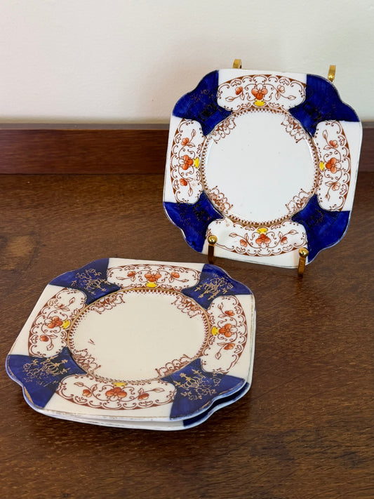 A stack of decorative ceramic plates with scalloped edges, featuring a white center, blue border sections, and intricate floral detailing in orange and gold, displayed on a wooden table with one plate propped up on a stand.