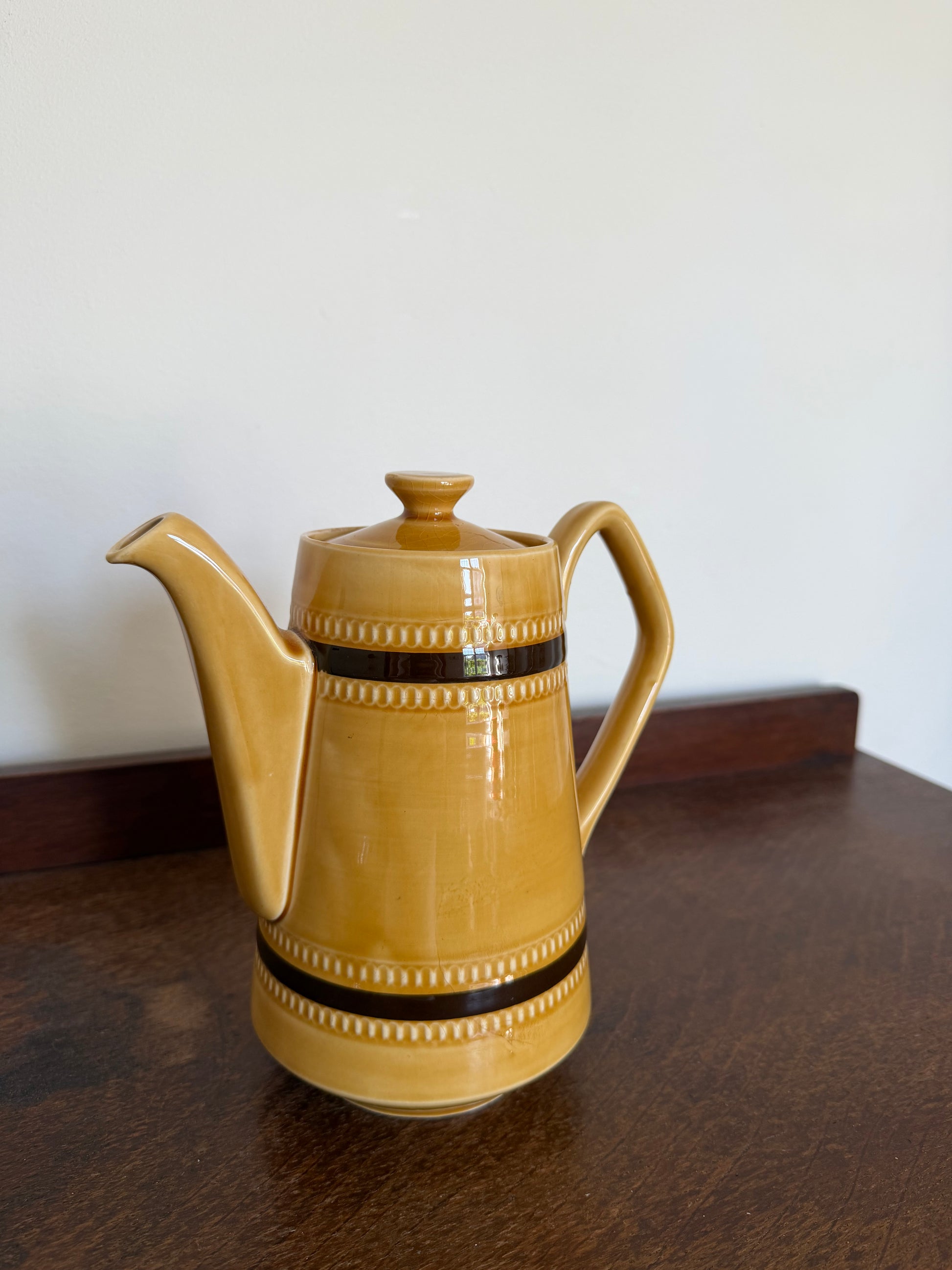 Vintage Sadler England coffee pot in a warm mustard yellow glaze with dark brown bands and raised beaded detailing. Tall cylindrical shape with a curved handle, long spout and matching lid, photographed on a dark wooden surface against a neutral wall.