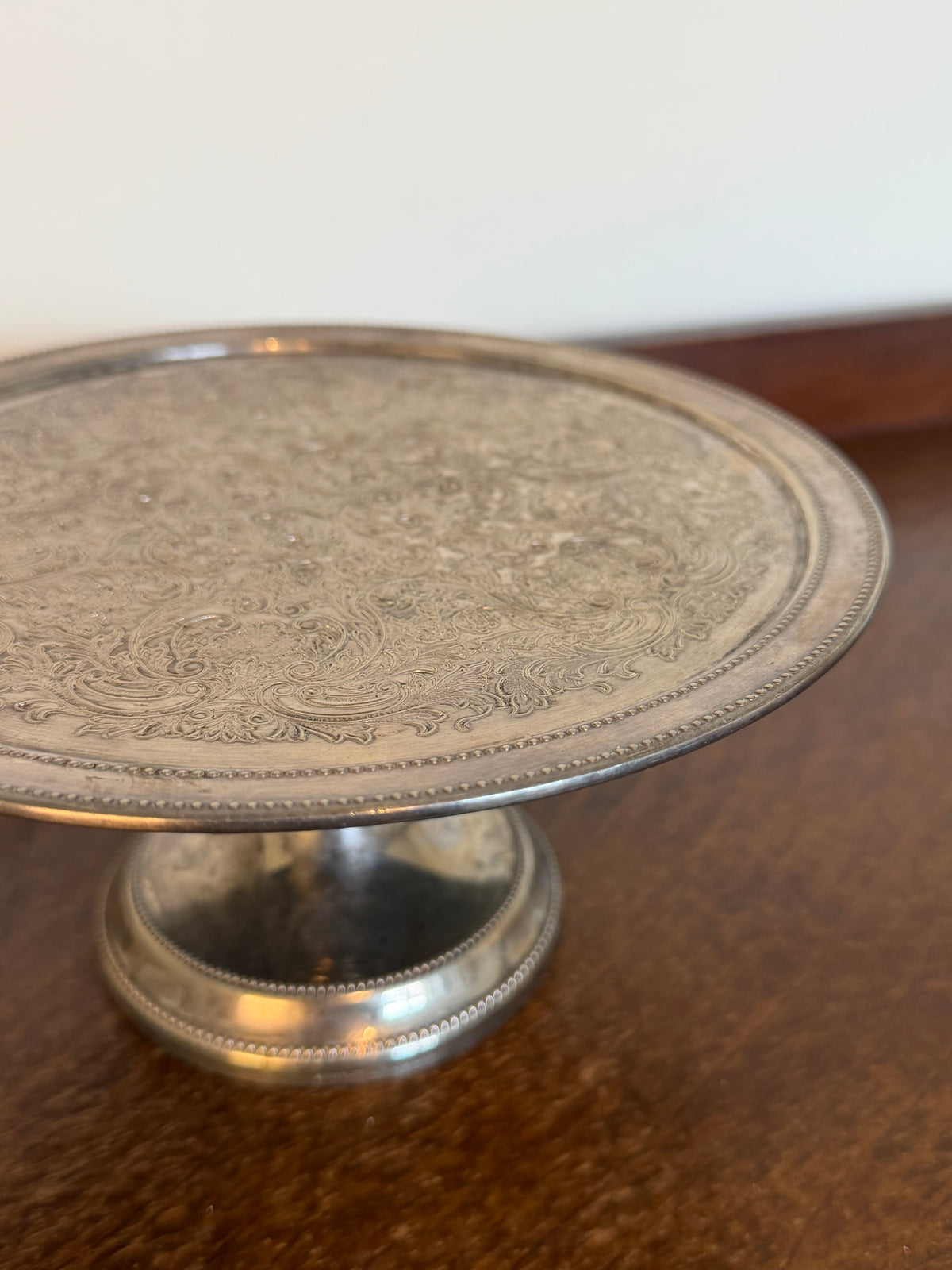 Vintage silver-plated pedestal cake stand with intricate floral scrollwork engraving, beaded edging and a decorative raised base, showing an aged tarnished silver finish.