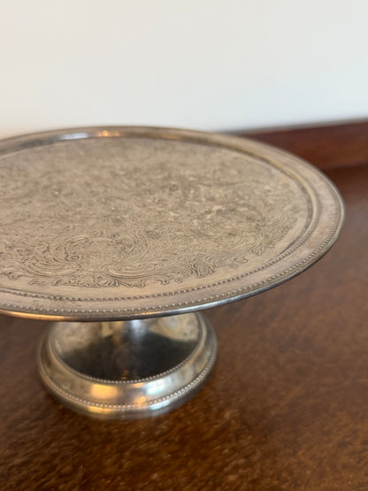 Vintage silver-plated pedestal cake stand with intricate floral scrollwork engraving, beaded edging and a decorative raised base, showing an aged tarnished silver finish.