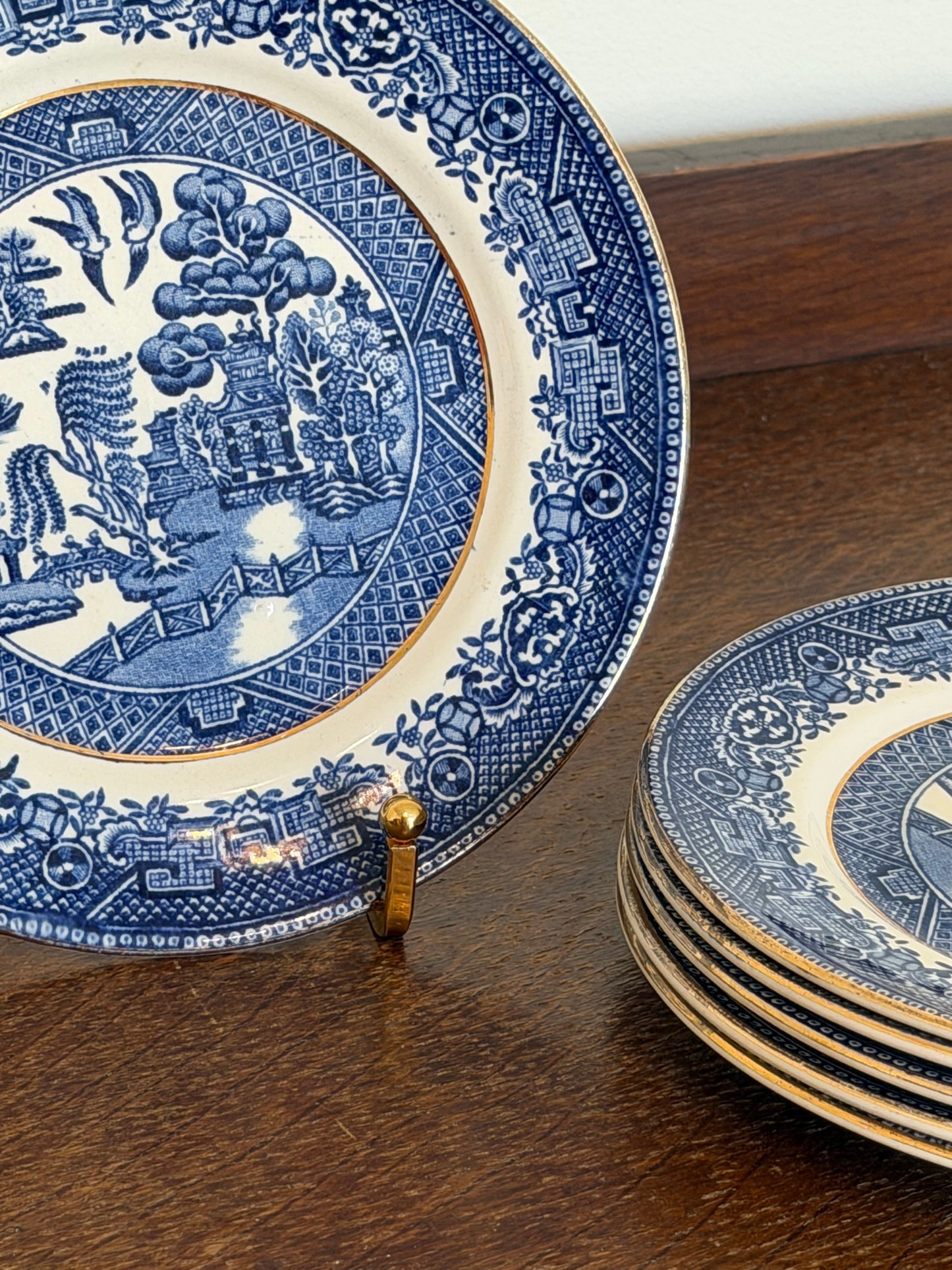 A close-up of a blue and white ceramic plate with a detailed landscape scene in the center, surrounded by a decorative border, resting on a dark wooden surface. A stack of similar plates is partially visible in the upper right.