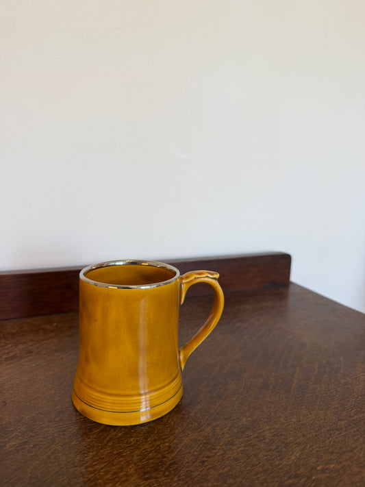 Vintage Wade ceramic tankard mug with a warm honey brown glazed finish, chunky curved handle and cylindrical shape, photographed against a neutral background. The underside shows the Wade England maker’s mark.