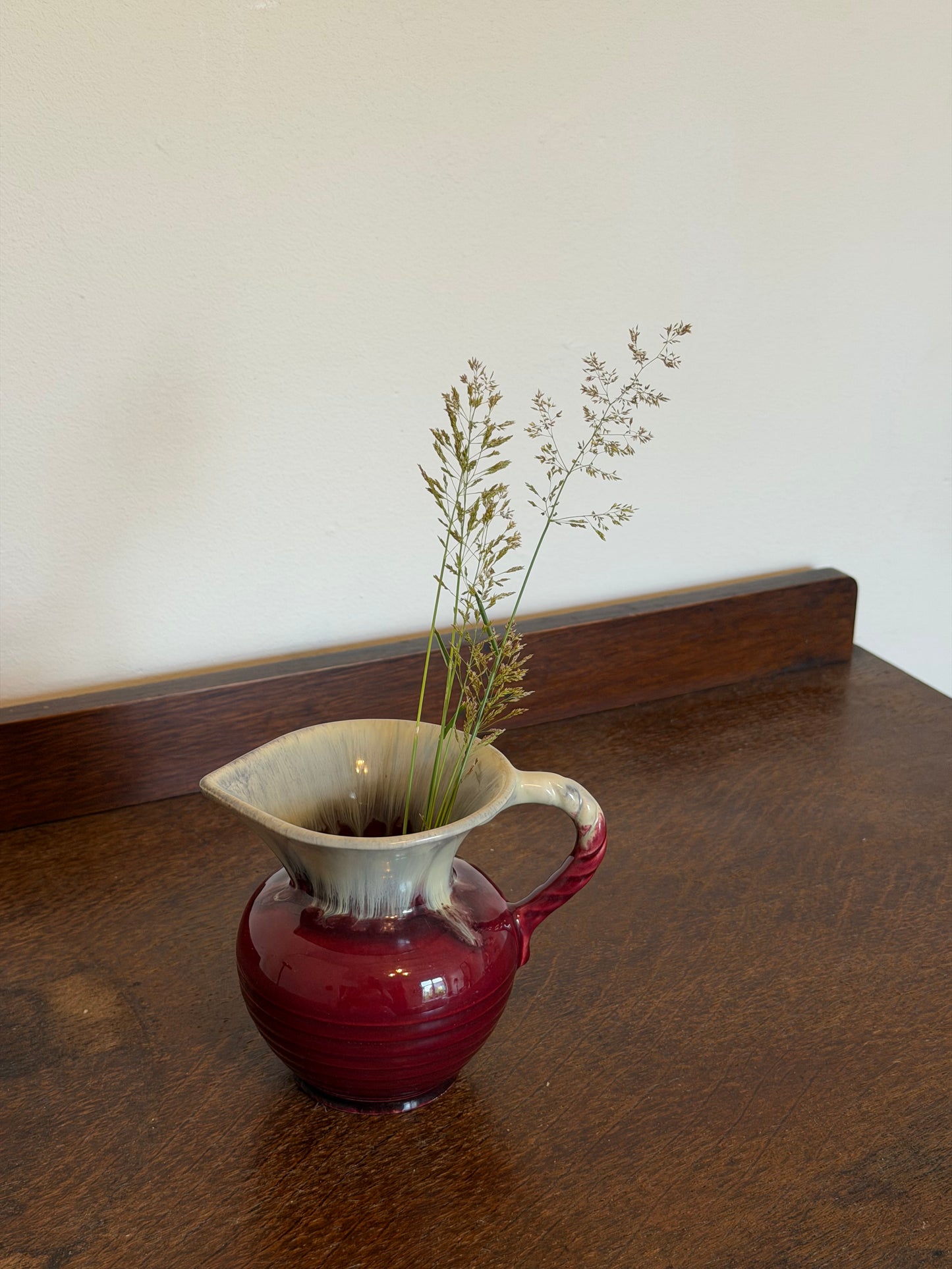 Small vintage West German ceramic jug with a rounded ribbed body in a glossy deep ruby red glaze and cream drip-glazed neck. The jug features a twisted handle, flared pouring lip and is photographed against an off-white wall on a dark wooden surface.