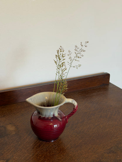 Small vintage West German ceramic jug with a rounded ribbed body in a glossy deep ruby red glaze and cream drip-glazed neck. The jug features a twisted handle, flared pouring lip and is photographed against an off-white wall on a dark wooden surface.